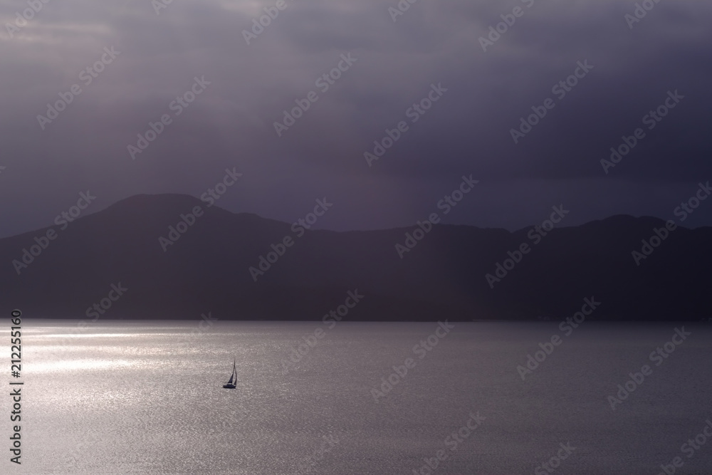 Fototapeta premium Barco veleiro solitário navengando no mar com montanhas ao fundo