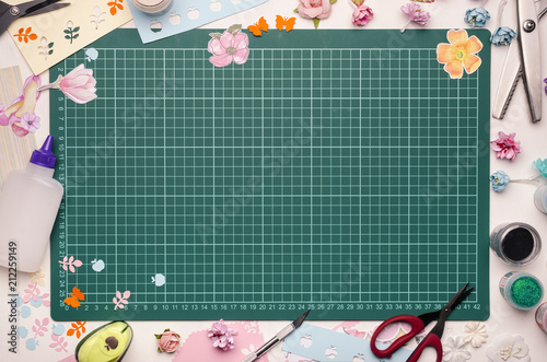 The cutting mat is surrounded by paper flowers, paper, tools and scrapbooking materials. Scrapbooking, top view