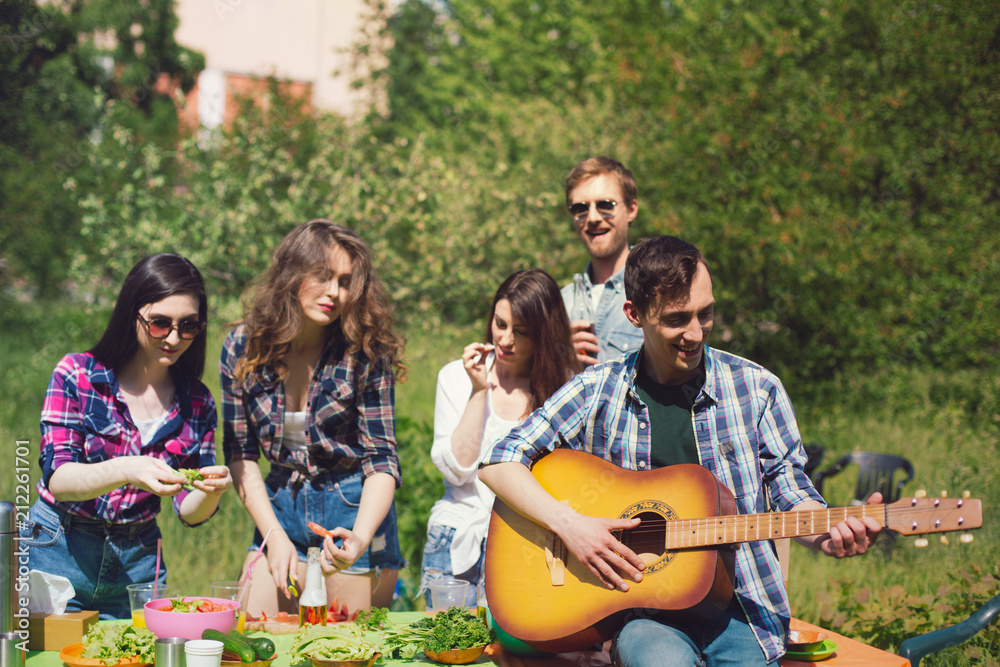 Fototapeta premium Company of friends enjoying time together on picnic. Guy playing guitar sitting on table out on picnic with his friends in background.