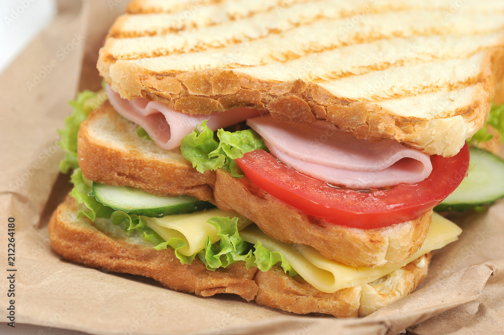 Sandwich with ham, cheese, fresh cucumbers and tomatoes. Slices of white bread are grilled. The sandwich lies on the kraft paper. Close-up. Macro photography.