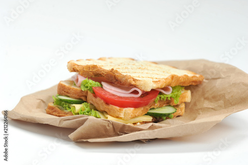 Sandwich with ham, cheese, lettuce, fresh cucumbers and tomatoes. Slices of white bread are grilled. The sandwich lies on the kraft paper. Close-up. White background.