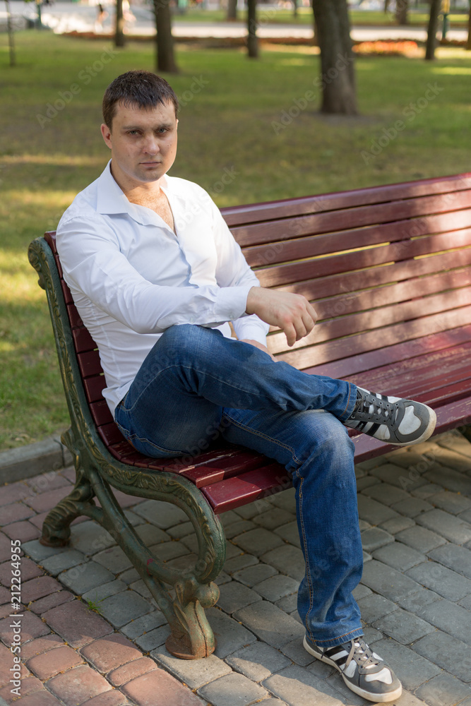 the guy is sitting on a bench in the Park. a man in a white shirt and jeans. young man. a man in a white shirt portrait of a man. business portrait. male portrai