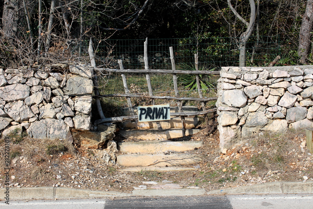 Abandoned garden with private sign on entrance surrounded with traditional stone wall and dried trees and branches in background Abandoned garden with private sign on entrance surrounded with traditional stone wall and dried trees and branches in background
