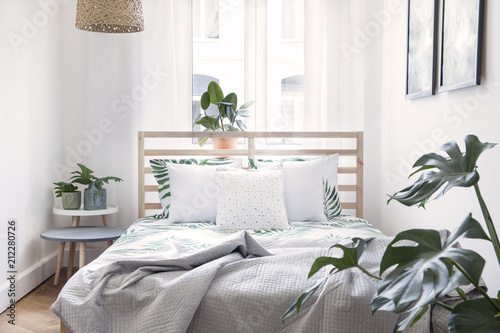 Modern scandinavian sunny bedroom with plants , floral pattern bedding and pilows. Space with white walls and brown wooden parquet. 