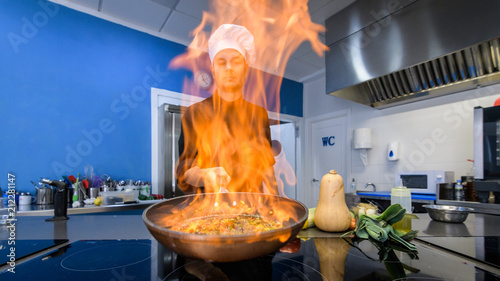 young chef flambe vegetables with fire in a frying pan, front view