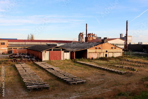 Abandoned wood furniture factory building made of old bricks with uncut grass, wood left for drying on air and other industrial building surrounding it at sunset