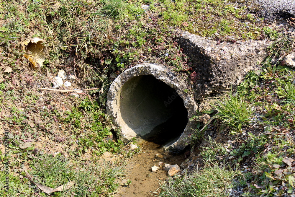 Damaged concrete drain pipe exit with running water and surrounded with dirt, stone, uncut grass, dried leaves and small damaged plastic pipe