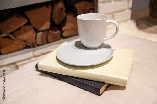 Coffee cup with saucer placed on books and pillow near firewood closeup - cosy home concept