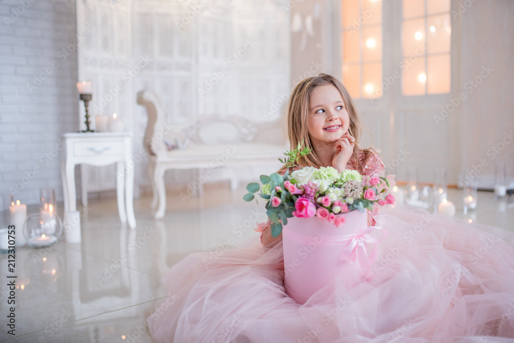 Little girl in pink dress holds box with roses sitting on the floor in a luxury room