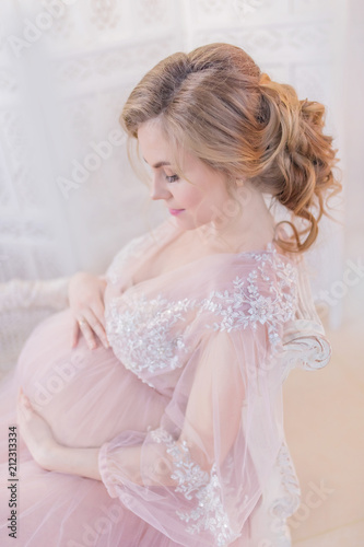 Stunning pregnant woman in pink dress rests on couch in a white room