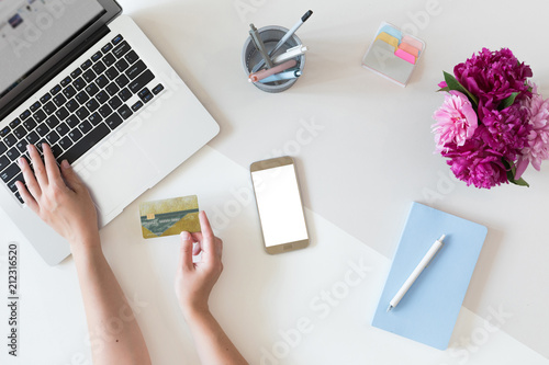 Top view of woman hands holding credit card, online shopping concept, workspace with laptop, mobile phone, flowers and notebook, flat lay.