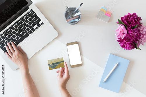 Top view of fashion office desk with credit card for online payment on laptop computer or mobile phone, flat lay on white background with cell phone and flowers