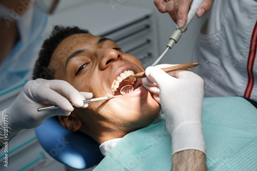 Dentist examining oral cavity of young African-American man working in in dental clinic with assistant. 