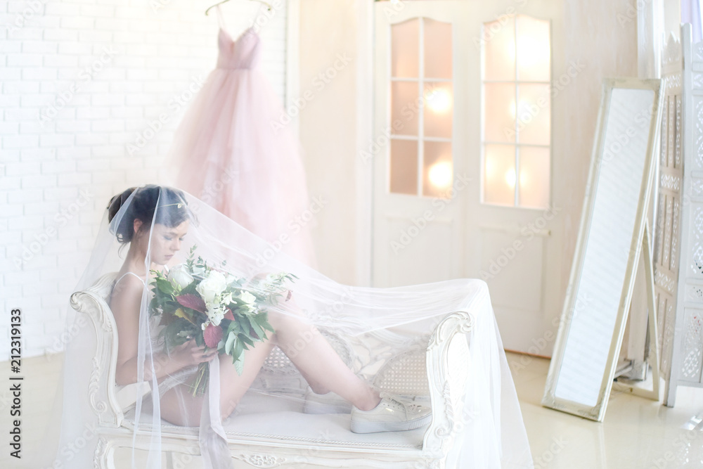 Bride in white lingerie and keds sits on a white couch under the veil Bride in white lingerie and keds sits on a white couch under the veil