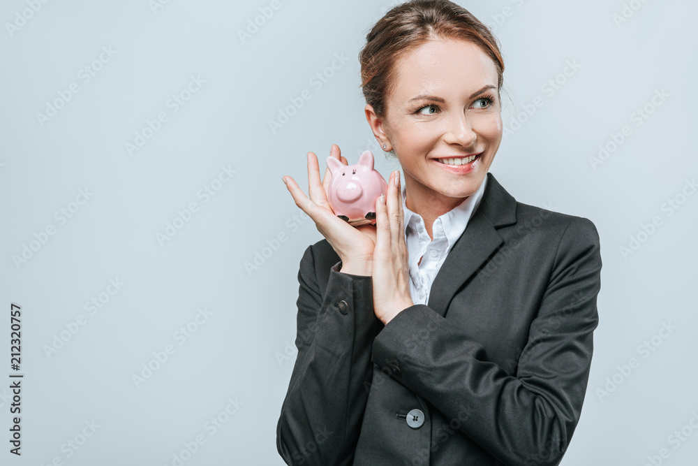 smiling financier holding piggy bank and looking away isolated on grey smiling financier holding piggy bank and looking away isolated on grey