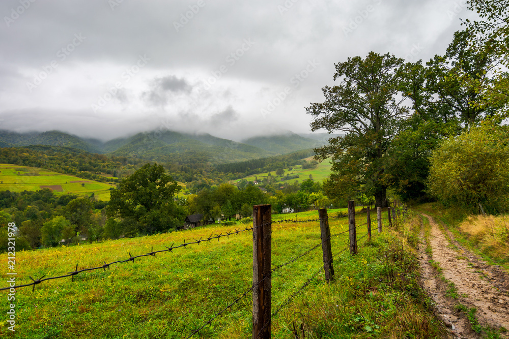 road along the fence in rural outskirts. pasture behind the barbwire. mountainous countryside on a dull day with overcast sky. village down in the valley. some old oak trees along the path