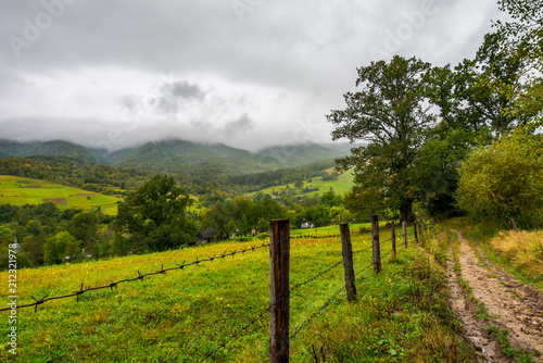 road along the fence in rural outskirts. pasture behind the barbwire. mountainous countryside on a dull day with overcast sky. village down in the valley. some old oak trees along the path © Pellinni