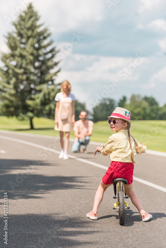 child in sunglasses riding bicycle while parents standing behind in park