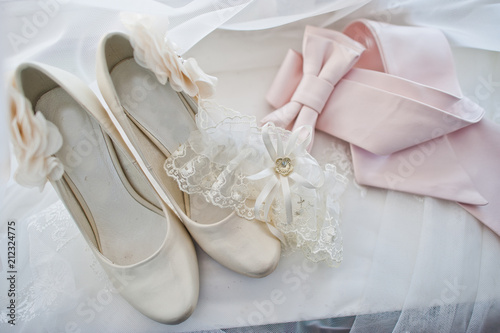 Close-up photo of bridal beige wedding heels, lace garter and pink ribbon on the white piece of fabric.