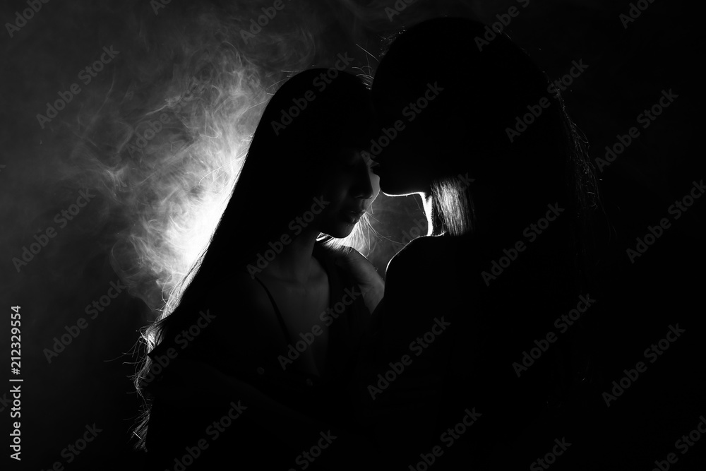 Silhouette of two sexy woman kissing holding in darkness through light and smoke