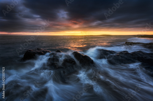 Long exposure of a dramatic and dark, coastal sunset with rocks low in the foreground. The sunlight is glowing across the horizon. The clouds are dark blue/purple and hanging low.