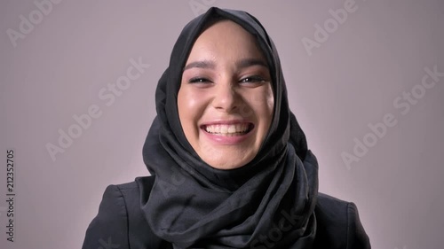 Young beautiful muslim girl in hijab is watching at camera, laughing, grey background, religious concept photo