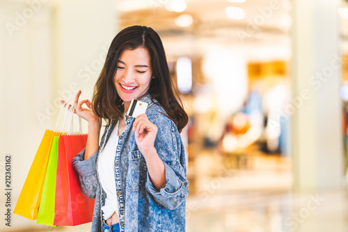 Happy beautiful Asian woman smile at credit card, hold shopping bags, copy space on shopping mall background. Shopaholic people, retail special offer price, holiday vacation activity lifestyle concept