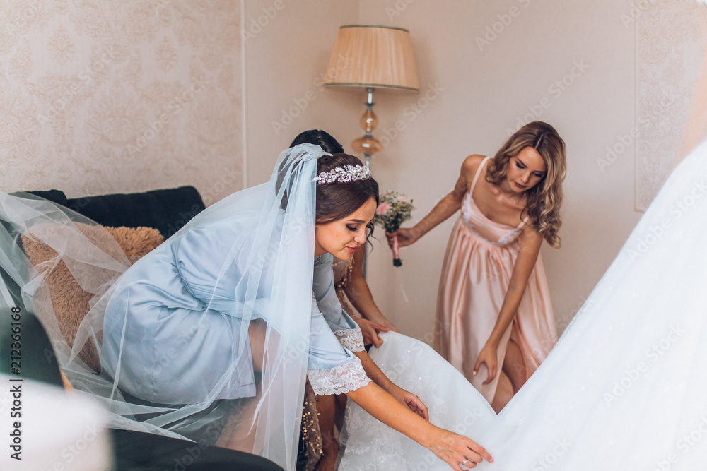 Bride on silk robe looking on her wedding dress against bridesmaids at room on morning day. - obrazy, fototapety, plakaty 