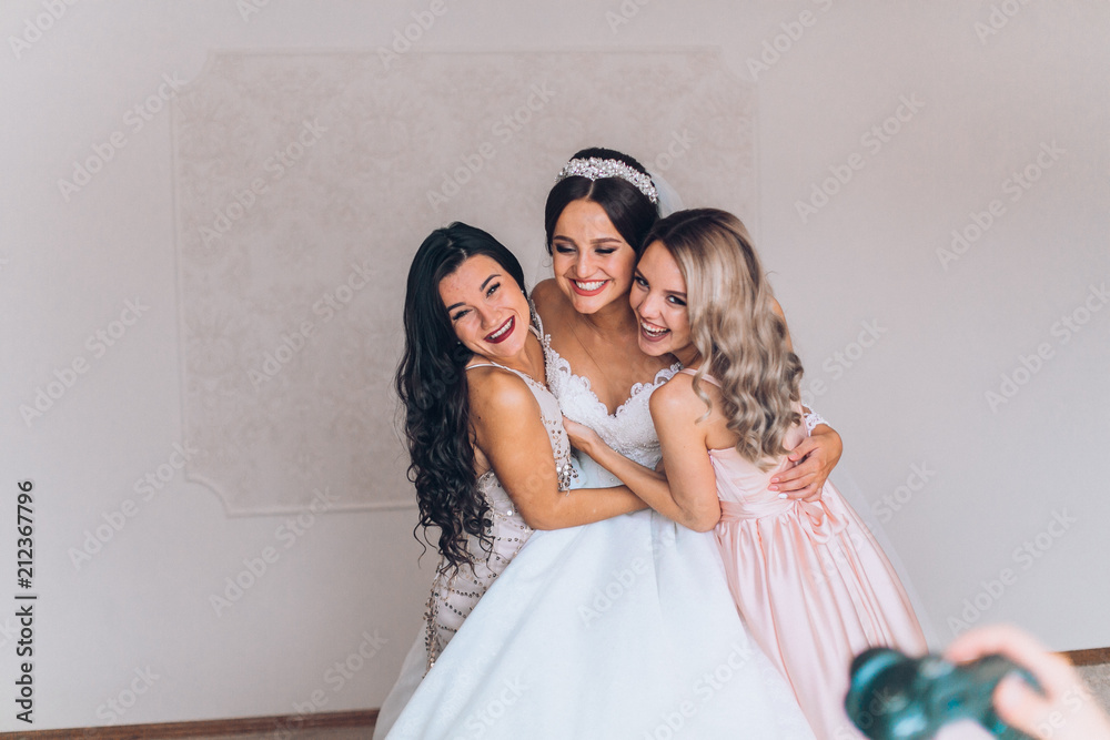 Happy bridesmaid hugging the bride in the bedroom in the morning. Before wedding ceremony. Bride preparing and wait groom. 