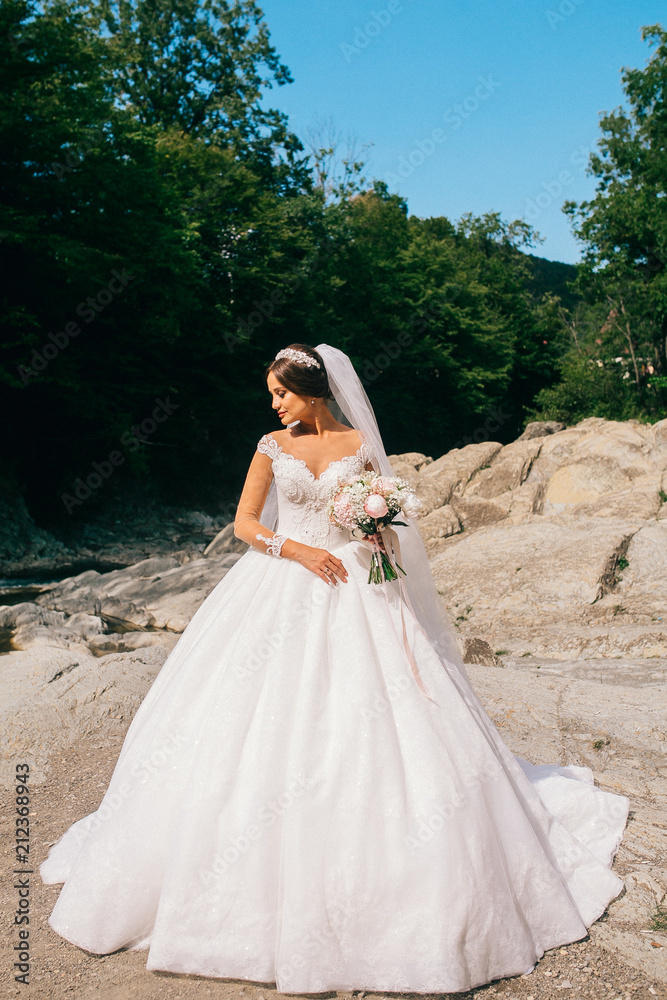 The bride in a wedding dress is standing on the mountain river bank with a bouquet of flowers. Wedding summer meadow. Young woman stay on stone.
