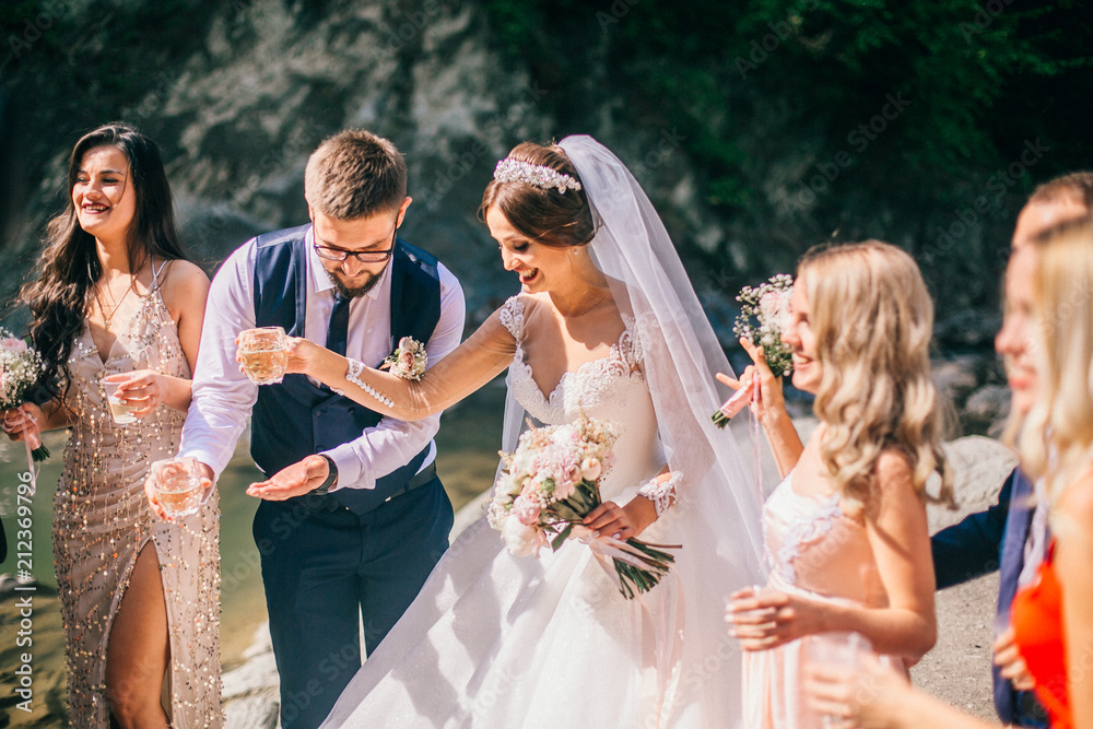 Wedding day bride and groom with bridesmaids and groomsmen posing in sunlight evening in mountains near river gorgeous wedding newlyweds couple with best friends. People drink champagne and hugging.