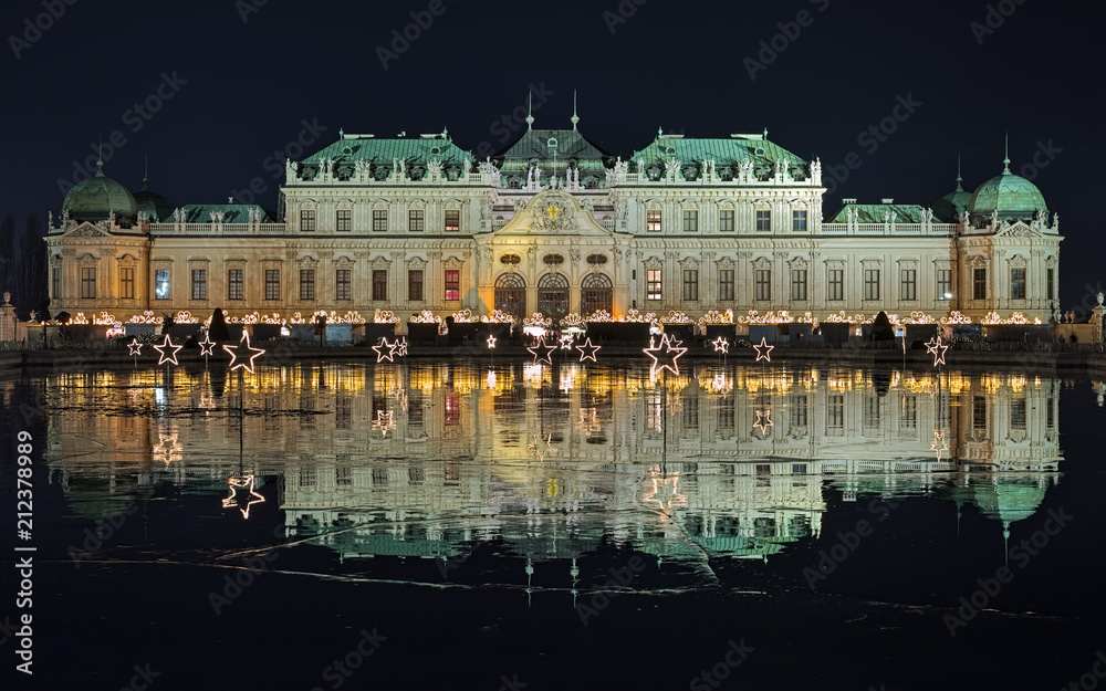 Upper Belvedere Palace with Christmas Village reflecting in the pond covered with wet ice in night, Vienna, Austria