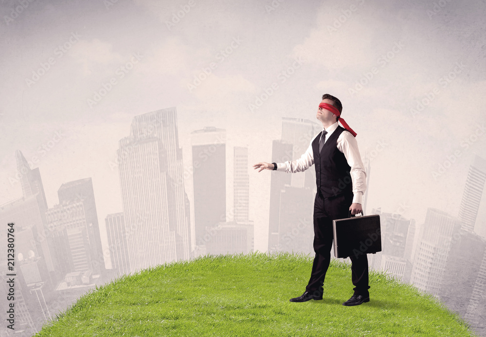 Young blindfolded businessman steps on a a patch of grass with a cloudy city in the background