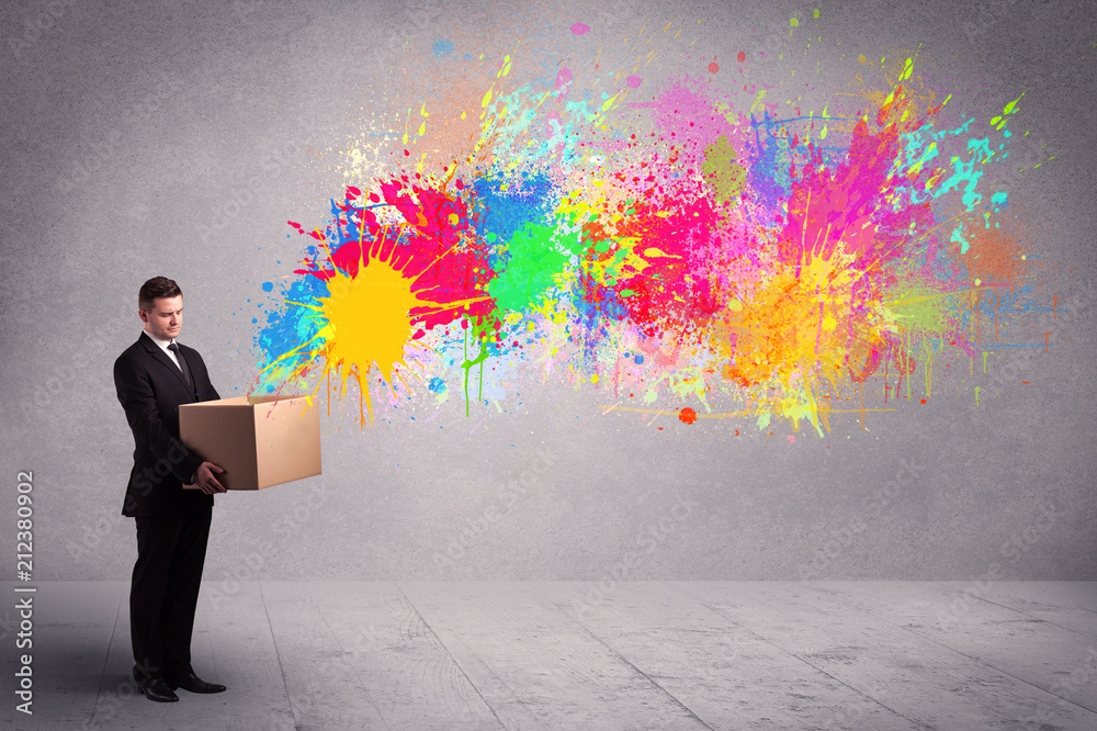 A young smiling business male holding a paperboard box with illustration of colourful spray paint splash on urban wall background concept. A young smiling business male holding a paperboard box with illustration of colourful spray paint splash on urban wall background concept.
