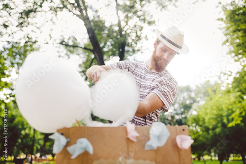 photo theme small business cooking sweets. A young man with a beard of a Caucasian trader in the hat the owner of the outlet makes candy floss, fairy floss or Cotton candy in the summer park