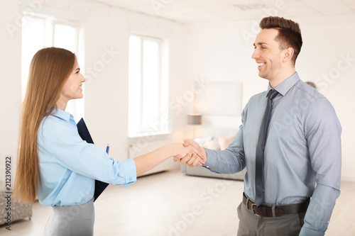 Beautiful real estate agent and young man shaking hands indoors