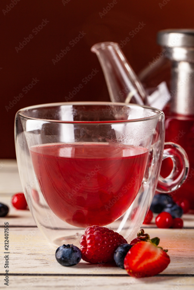 Mint tea with forest berries in a cup of double glass and a glass teapot on a wooden table, on a red background. Mint tea with forest berries in a cup of double glass and a glass teapot on a wooden table, on a red background.