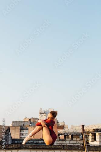 Young and graceful ballerina sitting on roof of city building at sunrise