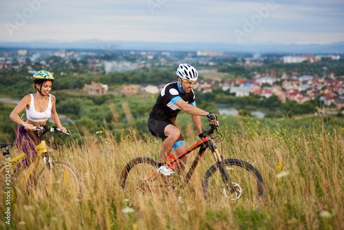 Guy and girl on bicycles ride at hill among the grass with wildflowers in the distance on a blurred background a town is visible. Guys in helmets and sportswear