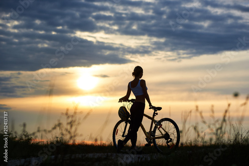 Silhouette of sporty cyclist, wearing sportswear and standing near her bicycle on trail. View of incognito woman enjoying nature and observing wonderful landscapes and amazing sunset. Back view