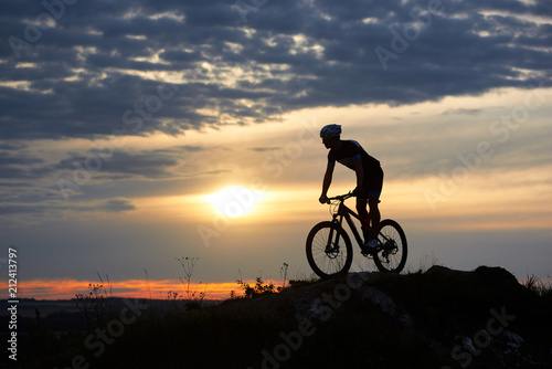 Silhouette of male cyclist wearing a helmet and sports clothes riding a mountain bike on rocks in the mountains against a beautiful sunset with a bright sun and sky in the clouds.