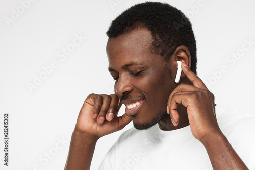 Young african american man wearing wireless earbuds and blank white t shirt, listening to his favorite musical album online, touching one earphone to control application photo