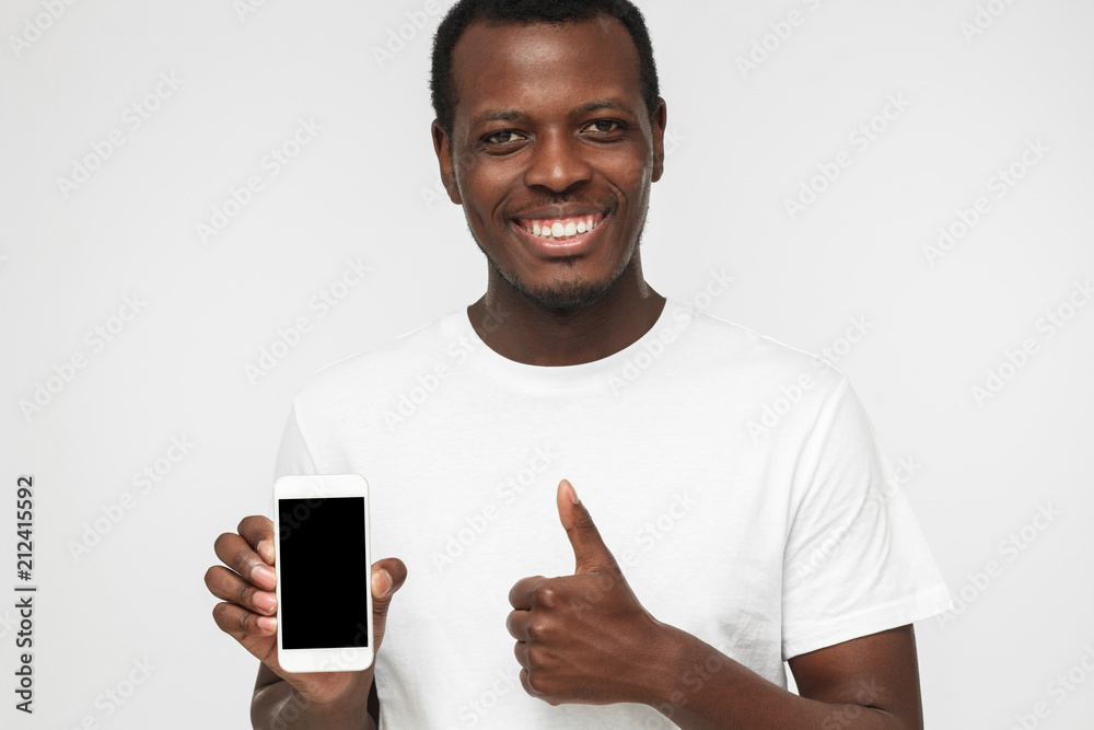 Closeup shot of young African American man isolated on white background wearing white t shirt, showing blank phone screen and thumb up willing to recommend product or service Closeup shot of young African American man isolated on white background wearing white t shirt, showing blank phone screen and thumb up willing to recommend product or service