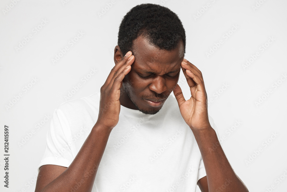 Young African American man in blank white t shirt isolated on gray background, feeling pain, touching temples with hands, feeling uneasy, tired and discontent Young African American man in blank white t shirt isolated on gray background, feeling pain, touching temples with hands, feeling uneasy, tired and discontent