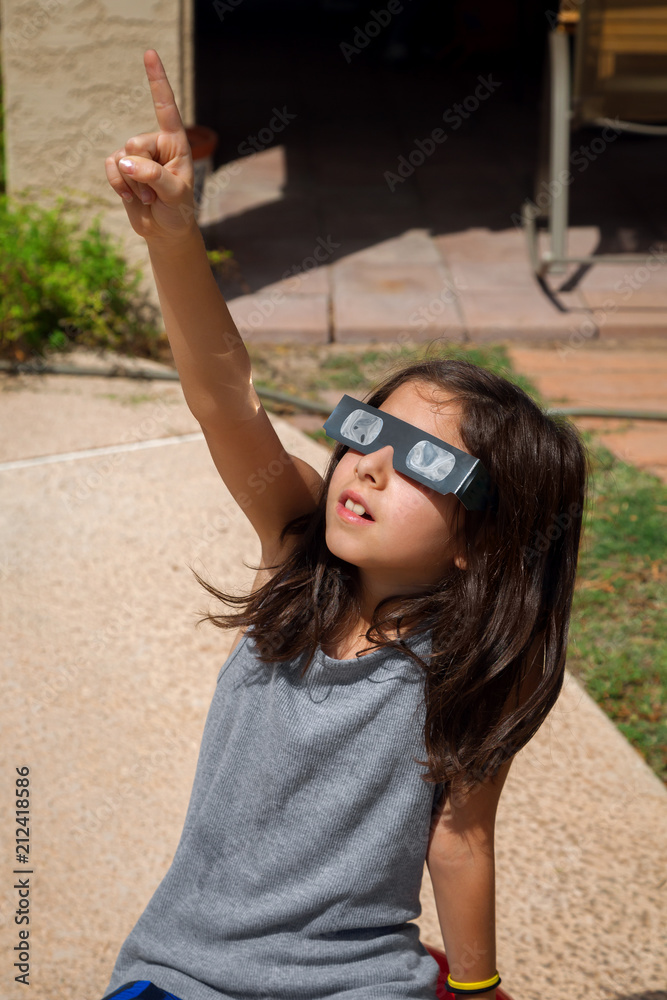 Fototapeta premium Young Girl Pointing To the Sky While Wearing Eclipse Glasses On a Bright Day