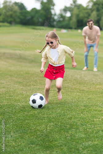 adorable child in sunglasses playing with soccer ball while father standing behind in oark