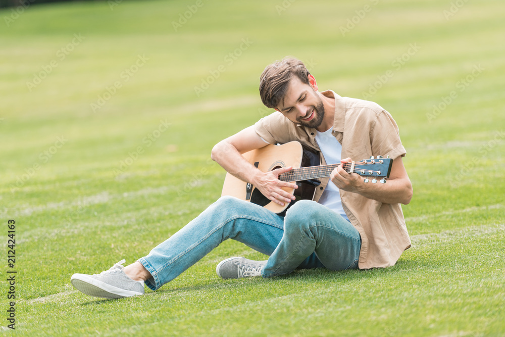 smiling young man sitting on lawn and playing acoustic guitar