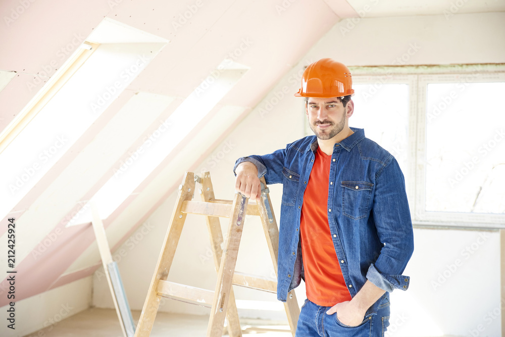 Interior home repair. Portrait of young repairman wearing hard hat while standing next to ladder at the construction site. 