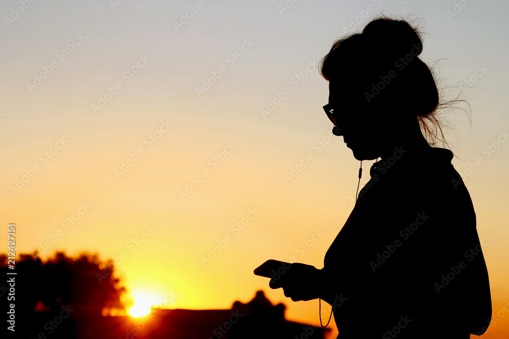 silhouette of woman with mobile phone and headphones on the background of sunset.