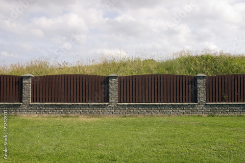 Long brown wooden fence from vertical pine planks and gray bricks.